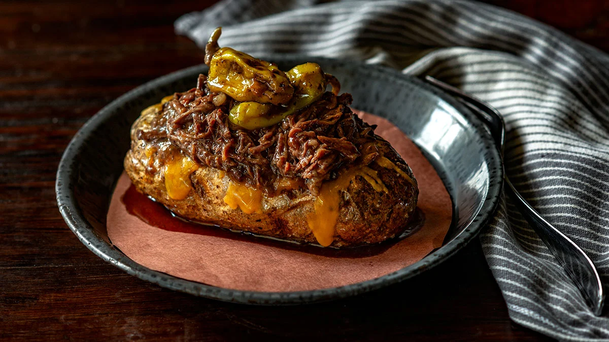 Baked potato topped with shredded beef, melted cheese, and sliced peppers on a black plate with a striped cloth in the background—perfect comfort food that brings the warmth of Sunday Suppers and home-cooked meals to your table.
