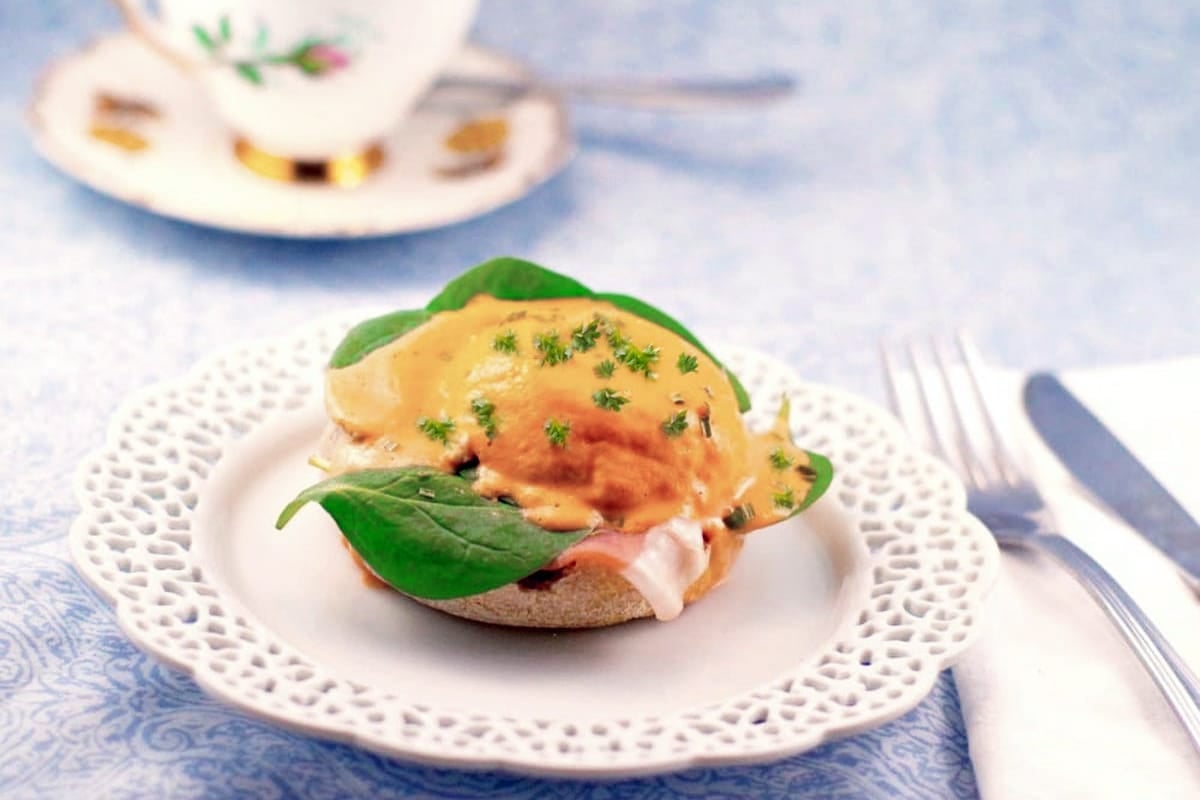Eggs Benedict with spinach, ham, and hollandaise sauce on an English muffin, topped with a touch of roasted peppers for extra flavor, served on a decorative white plate with a fork, knife, and teacup in the background. Perfect inspiration for those seeking new roasted peppers recipes.