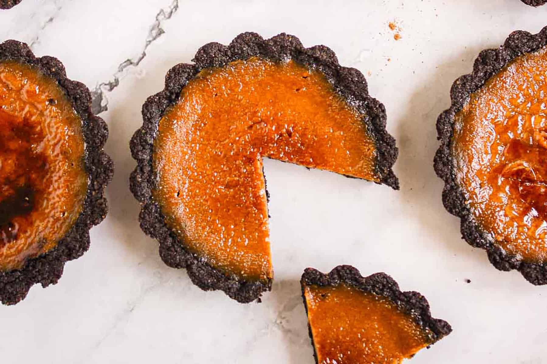 Three small round tarts with dark chocolate crusts and caramelized tops are on a marble surface; one tart has a slice cut out and placed nearby. Perfect for those seeking new pumpkin dishes, these treats are a delicious addition to your autumn recipes collection.