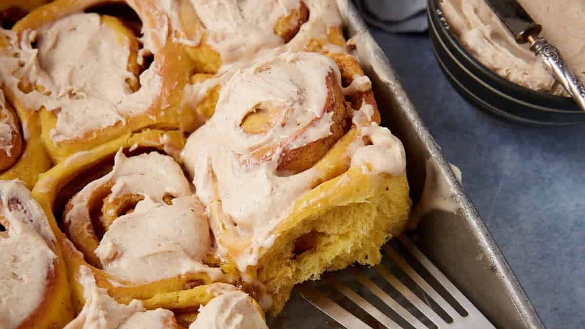 A baking tray of frosted cinnamon rolls, with one roll partially removed using a metal spatula, makes a perfect treat for autumn. A bowl of extra frosting and a butter knife are nearby, offering inspiration for new pumpkin recipes to enjoy this cozy season.