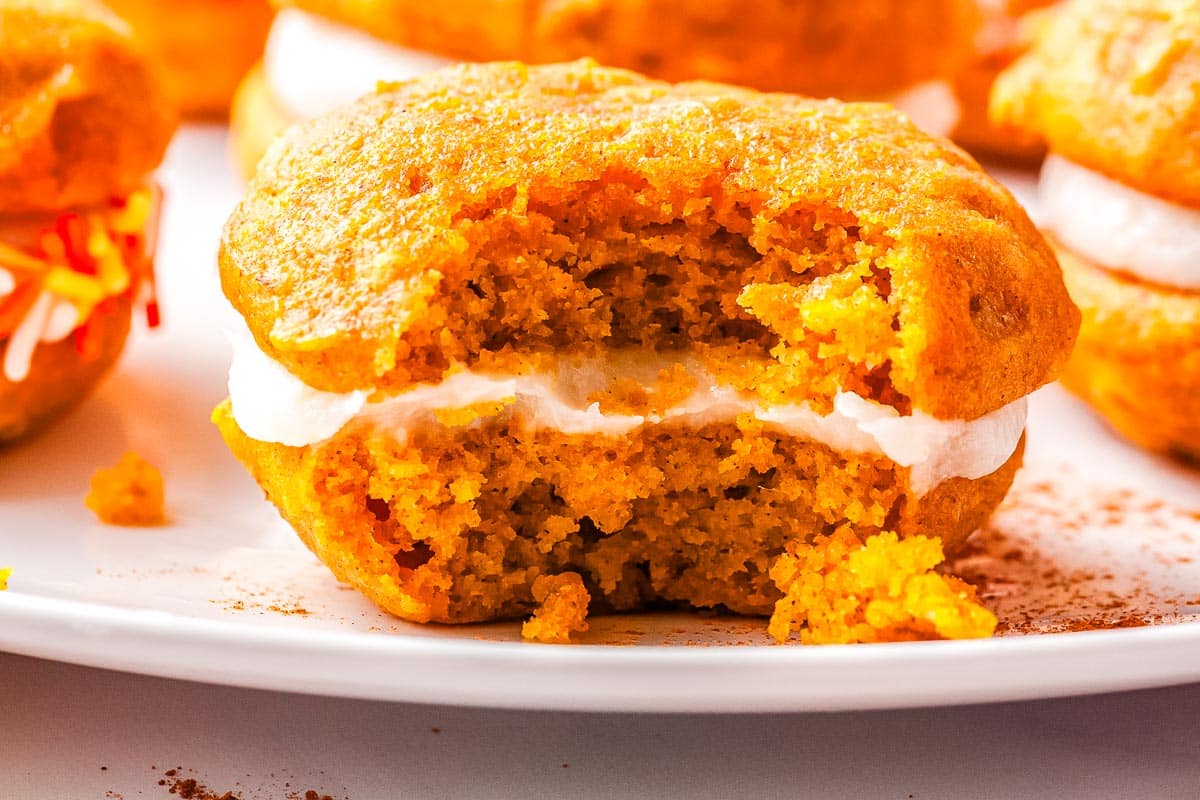 A pumpkin whoopie pie with a bite taken out, showing its creamy white filling, sits on a white plate with crumbs around it—an inviting treat that’s perfect for autumn desserts.