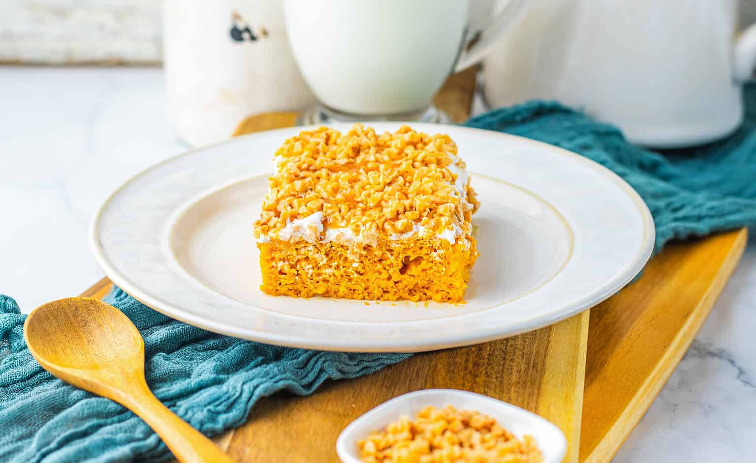 A square slice of yellow cake topped with chopped nuts sits on a white plate, with a wooden spoon and extra nuts nearby on a blue cloth and wooden board—an inviting treat perfect for autumn recipes or as inspiration for new pumpkin dishes.