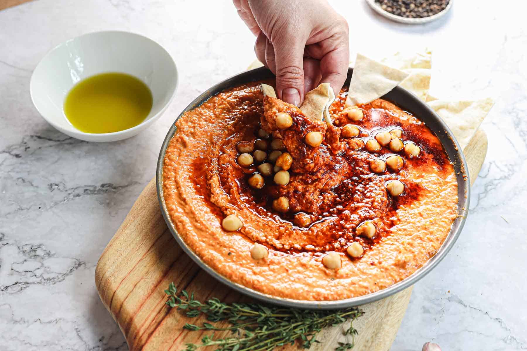 A hand dips pita bread into a bowl of hummus topped with chickpeas and spices, while a vibrant jar of roasted peppers adds color to the wooden board alongside a bowl of olive oil and fresh thyme.