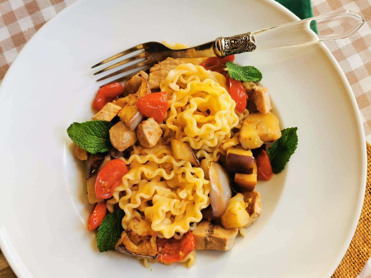 Plate of pasta with grilled chicken, cherry tomatoes, eggplant, zucchini, and fresh mint leaves, served alongside a fork on a white plate and set atop a checkered tablecloth. For those who enjoy exploring new flavors beyond poultry, this dish pairs beautifully with easy fish recipes for a delightful meal experience.