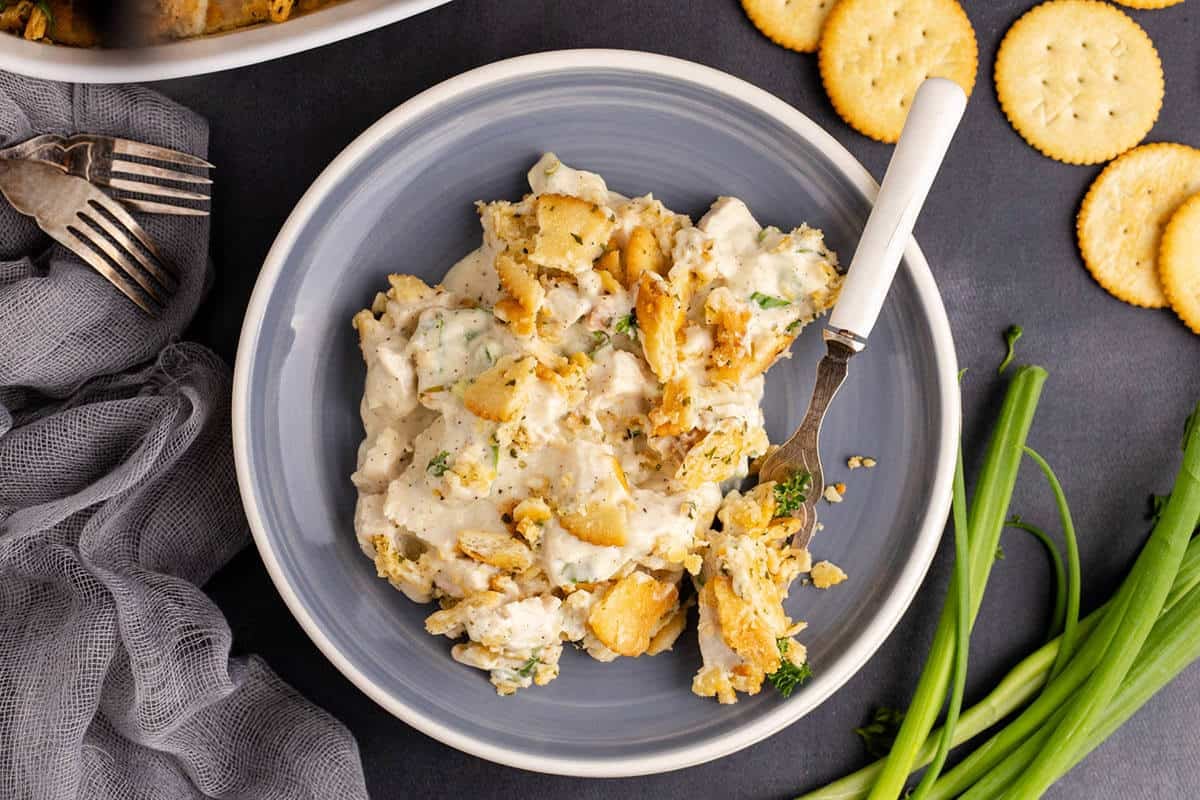 A plate of creamy chicken dinner casserole with chunks of crackers and herbs, served with a fork. Green onions and round crackers are beside the plate on a dark surface.