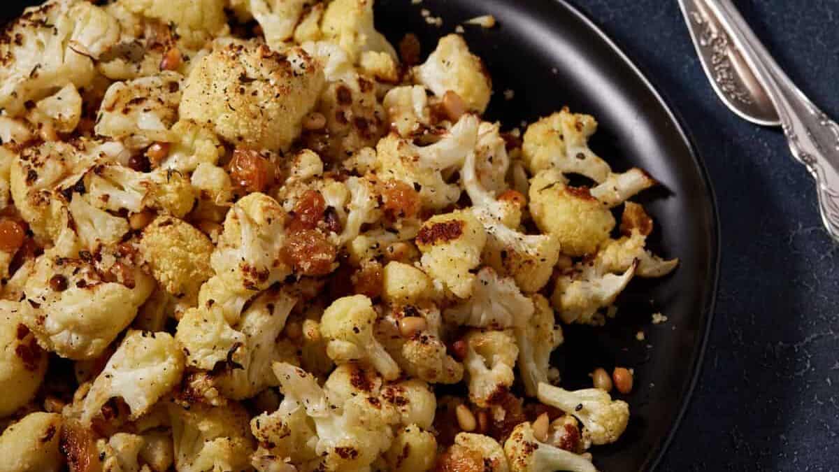 Roasted cauliflower florets with toasted pine nuts and golden raisins on a black plate, perfect for those seeking delicious roast vegetable recipes.