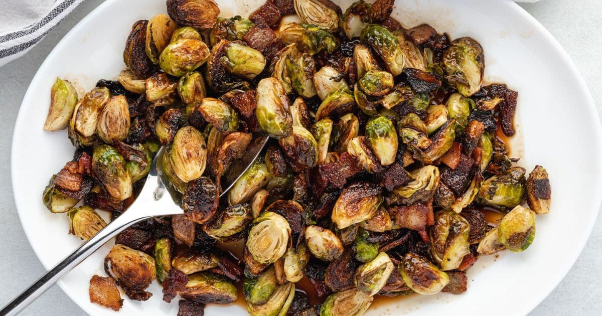 A white plate filled with roasted Brussels sprouts and crispy bacon, served as one of the healthy side dishes, with a serving spoon resting on the plate.