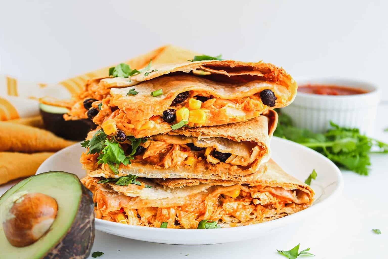 Three quesadilla wedges stacked on a white plate, filled with shredded chicken, black beans, corn, cheese, and garnished with cilantro—perfect for easy family dinners or kid-friendly meals. Sliced avocado and a bowl of salsa sit in the background, making this a delicious addition to your collection of family dinner recipes.