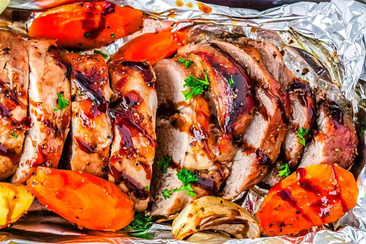 Sliced roasted pork tenderloin with glaze, garnished with parsley, served with roasted carrots and onions on aluminum foil—perfect for easy recipes and delicious weeknight dinners.