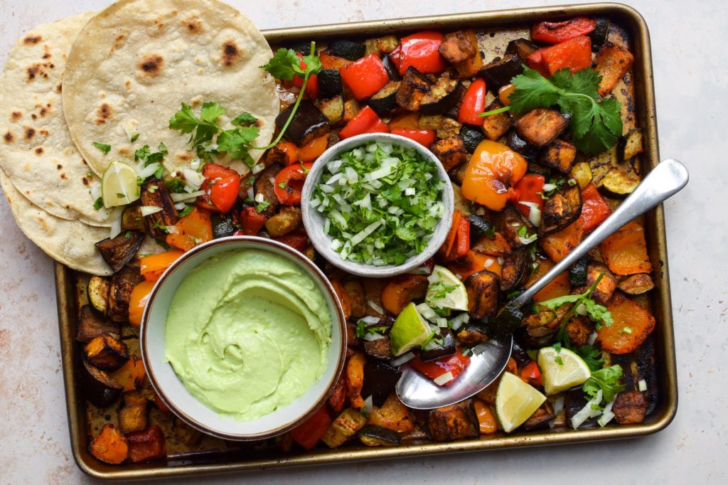 Sheet pan with roasted vegetables, flour tortillas, avocado dip, salsa with chopped cilantro, lime wedges, and a serving spoon arranged on a baking tray—perfect for easy sheet pan meals that make weeknight dinners effortless and delicious.