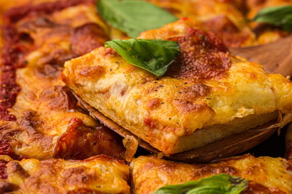 A close-up of a rectangular slice of cheesy, thick-crust pizza with pepperoni and fresh basil, being lifted from the rest of the pizza—an irresistible sheet pan meal that’s perfect for easy dinners or weeknight meals.