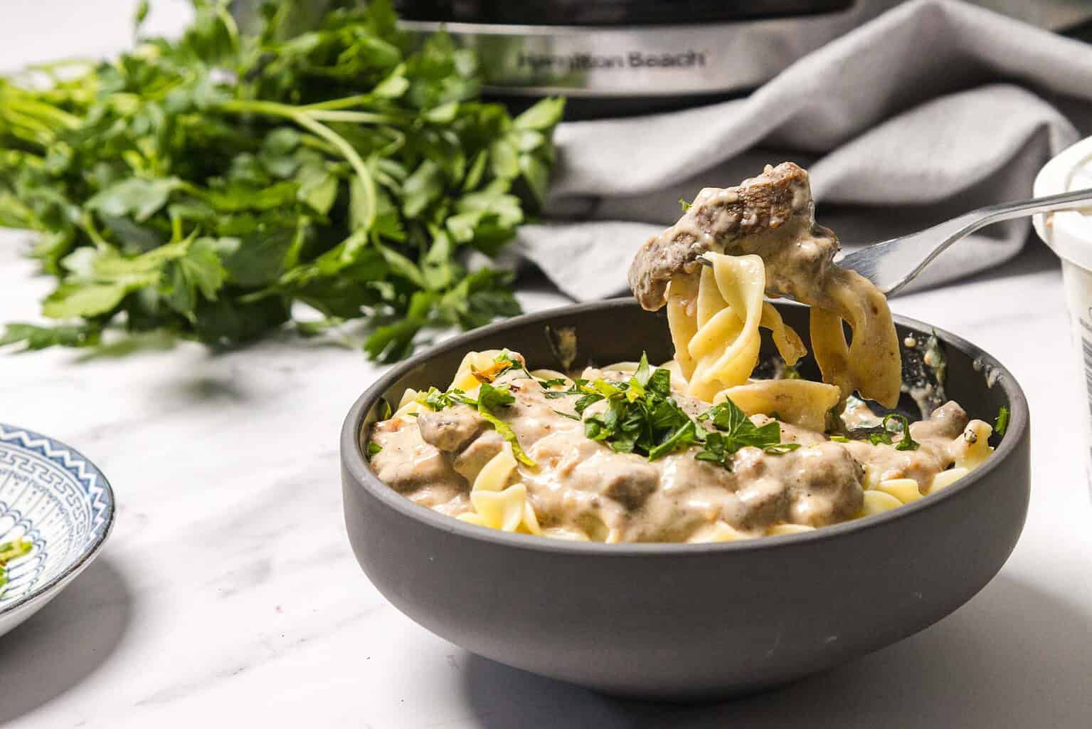 A bowl of pasta with creamy sauce and ground meat, topped with chopped parsley, sits on a marble surface next to fresh herbs—a true staple among essential recipes.