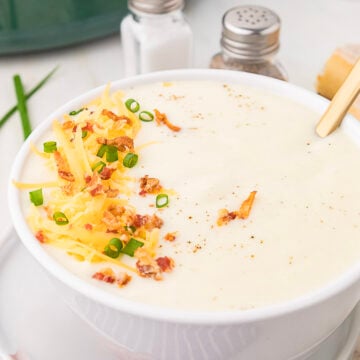 A bowl of creamy slow cooker cauliflower potato soup topped with shredded cheddar cheese, bacon bits, and chopped green onions.