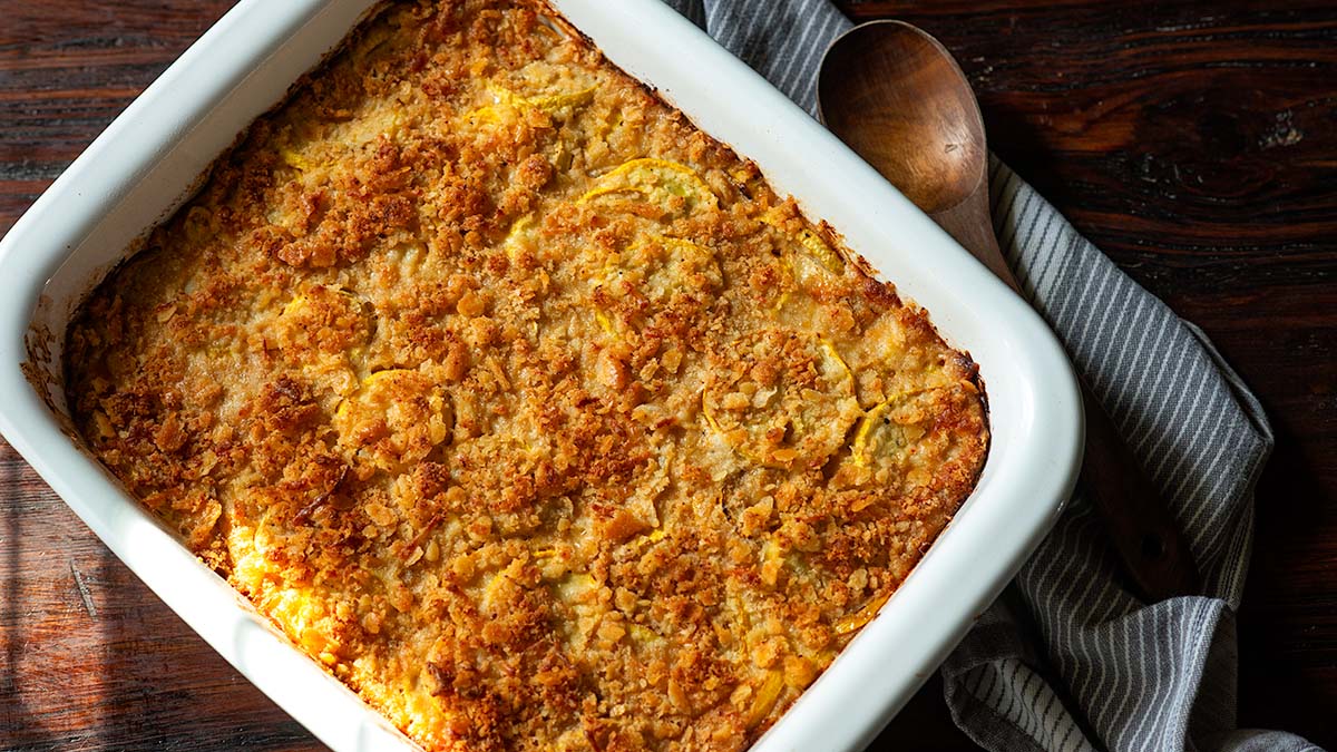 A baked yellow squash casserole with a golden, crispy topping in a white dish sits on a wooden table next to a striped cloth and a wooden spoon—a flavor-packed addition to your favorite side dishes and recipes.