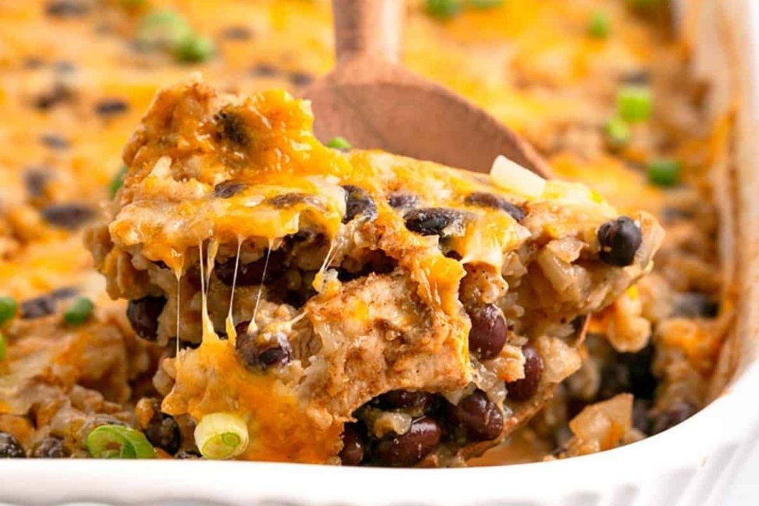 A wooden spoon lifts a cheesy black bean and rice casserole with rotisserie chicken from a white baking dish, topped with melted cheese and green onions.