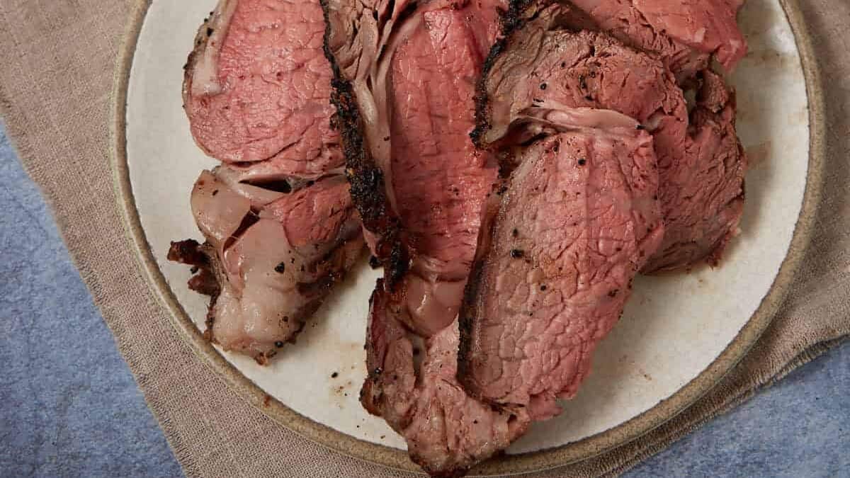 Sliced medium-rare roast beef on a round white plate, placed on a tan cloth with a blue surface underneath—a classic comfort food perfect for home cooking and hearty Sunday suppers.