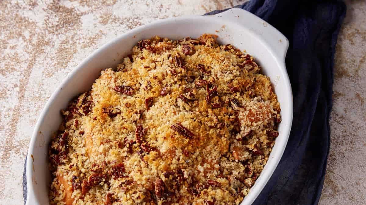 Oval white baking dish filled with a golden brown pecan and crumb-topped casserole, resting on a dark cloth—perfect for Thanksgiving and sure to become one of your favorite side dish recipes.
