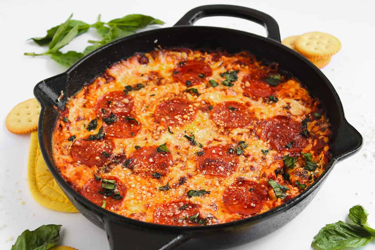 A cast iron skillet filled with baked dip topped with melted cheese, pepperoni slices, and fresh basil, surrounded by crackers and basil leaves—the perfect addition to your collection of sweet and savory recipes.