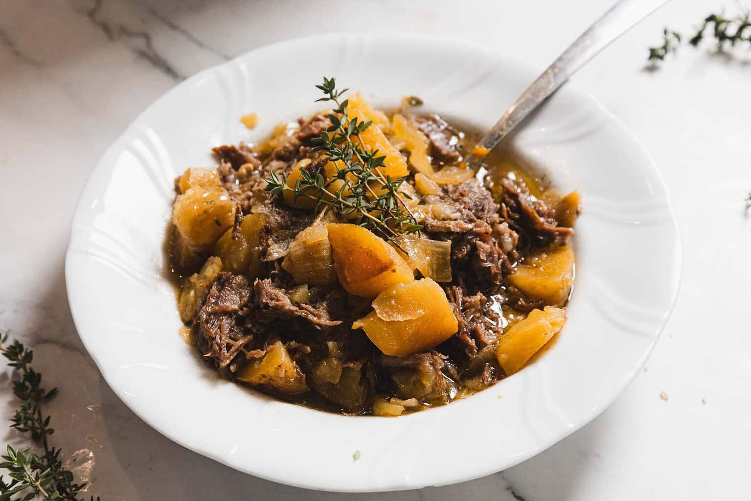 White bowl filled with beef stew, chunks of meat, potatoes, and vegetables, garnished with thyme. Try this recipe with a hint of maple syrup for added richness. Spoon rests on marble surface.