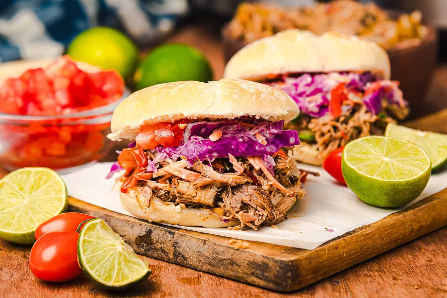 Two pulled pork sandwiches with red cabbage slaw and sliced tomatoes are served on a wooden board with lime halves and cherry tomatoes beside them—a satisfying nod to Tex Mex recipes perfect for hearty dinner recipes.