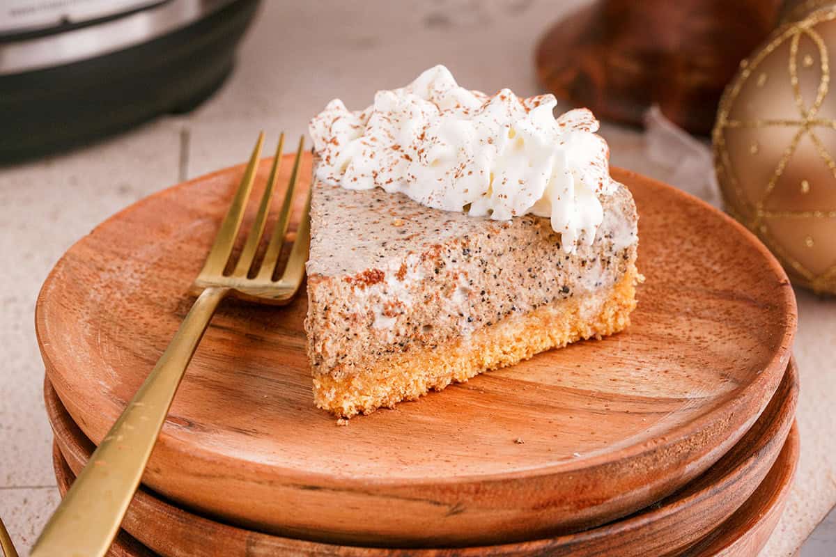 A slice of speckled cheesecake with whipped cream on top sits on a wooden plate with a gold fork beside it.
