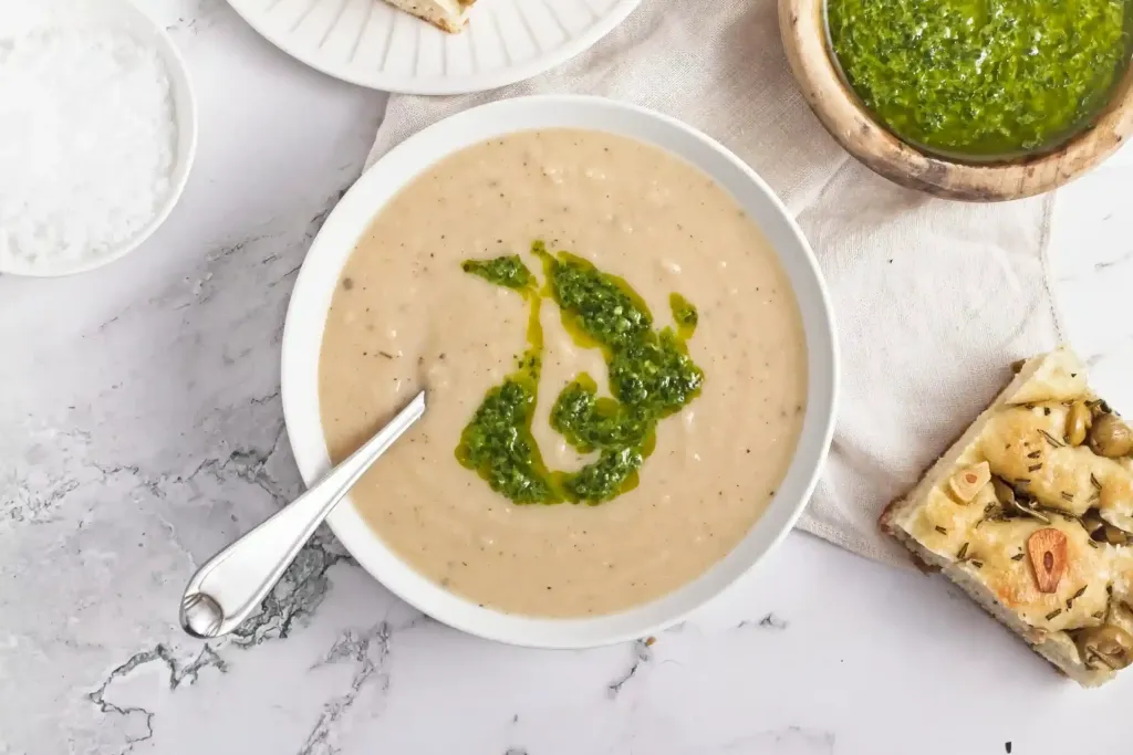 Bowl of creamy soup with green pesto drizzle, served with a spoon. Perfect for those seeking comforting soup options, this dish is accompanied by focaccia and a small bowl of pesto placed nearby on a marble surface—a delicious addition to your collection of bean recipes.