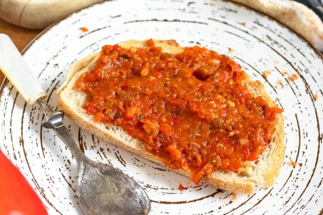 A slice of bread topped with a vibrant red vegetable spread, made from roasted peppers, sits on a white plate with a spoon next to it.