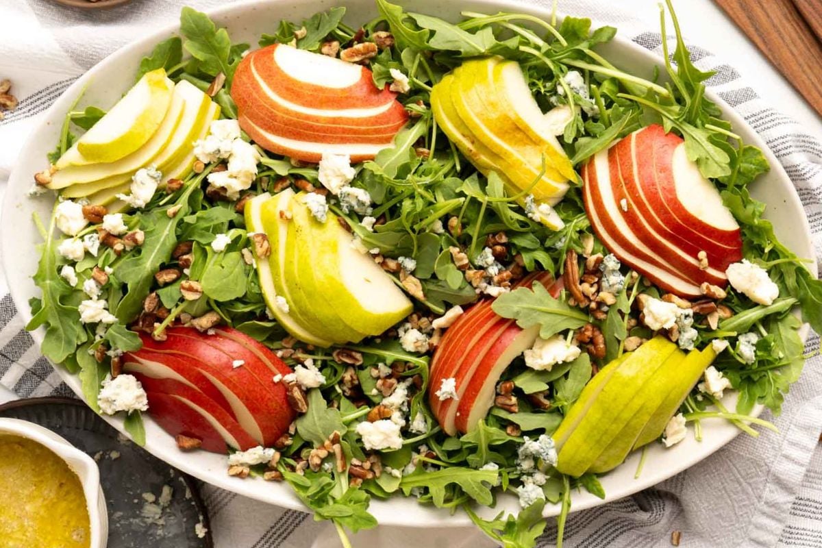 A salad with arugula, sliced red and green apples, pears, crumbled cheese, and chopped nuts on a white platter is the perfect addition to your flavor-packed recipes for a refreshing twist on your dinner routine.