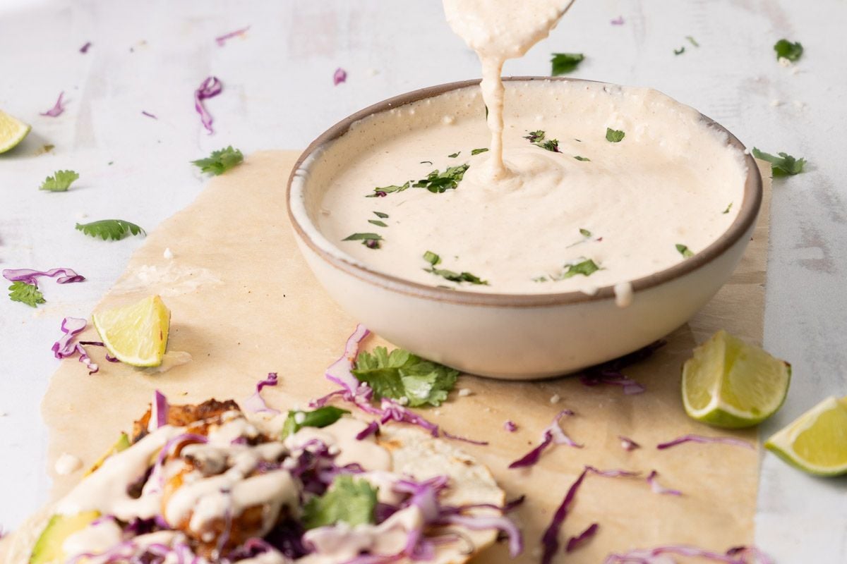 A bowl of creamy Tex Mex sauce with herbs is shown, with the sauce being drizzled. Shredded cabbage, lime wedges, and a taco topped with the flavorful sauce are nearby on parchment paper, making it perfect for your favorite dinner recipes or any Tex Mex meal.