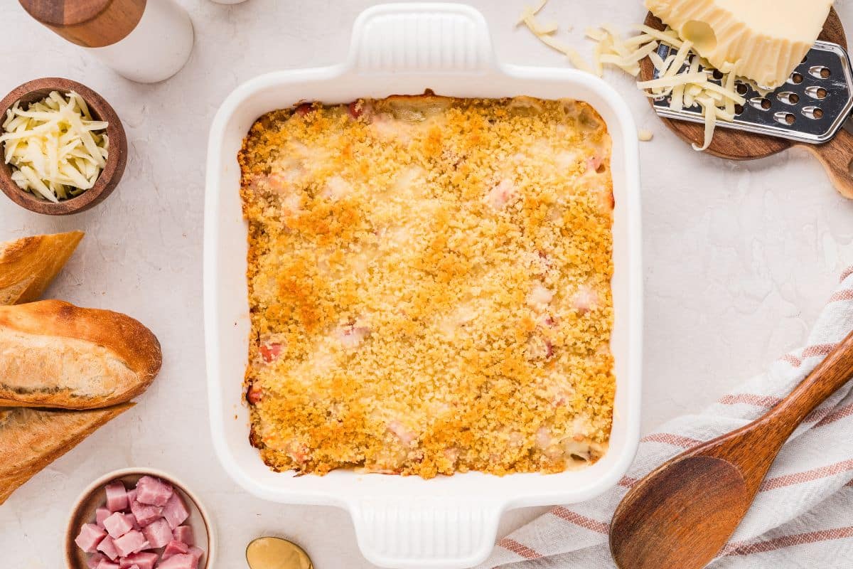 A baked casserole with a golden breadcrumb topping in a white dish, surrounded by bread, cheese, diced ham, and kitchen utensils on a light surface—perfect inspiration for those seeking unique casserole ideas or delicious new casserole recipes.