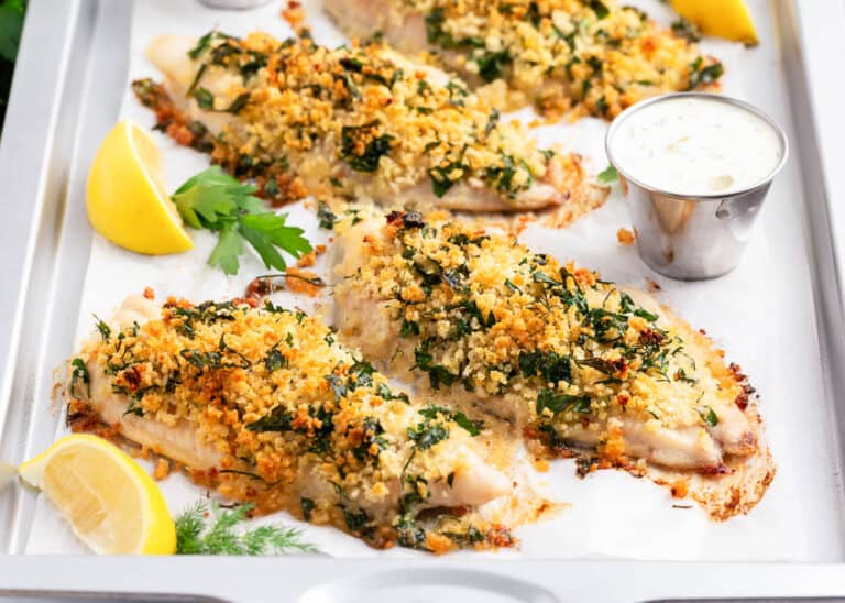 Baked fish fillets topped with a golden breadcrumb and herb crust, served with lemon wedges, fresh herbs, and a cup of creamy dipping sauce on a white tray—this is one of the best fish dishes for any occasion. Perfect for those seeking easy fish recipes that don’t compromise on flavor or presentation.