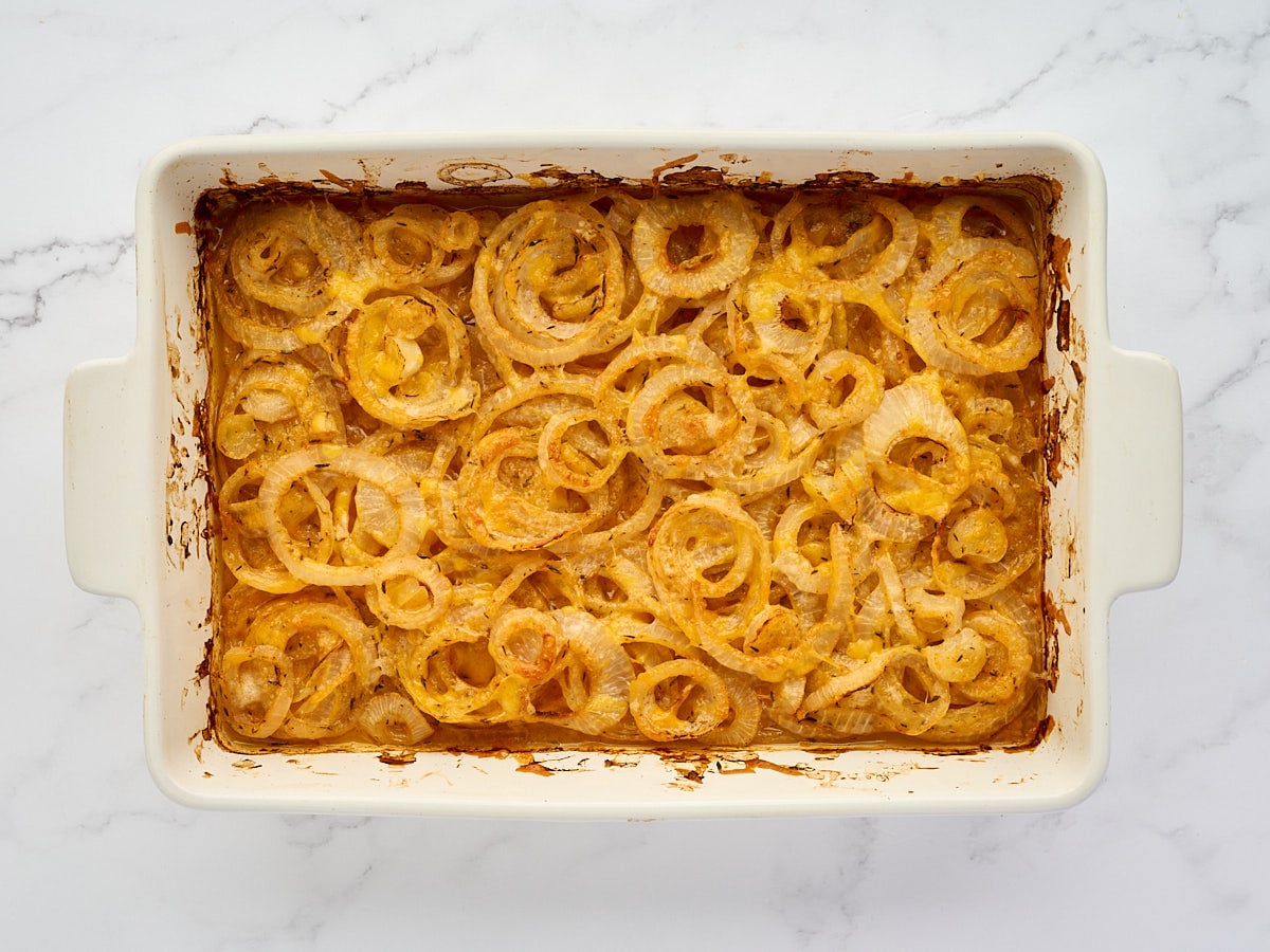 Rectangular white baking dish filled with baked sliced onions in a creamy, browned sauce, on a white marble surface.