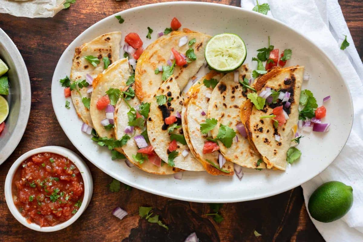 Five folded tacos filled with vegetables, topped with cilantro, diced tomato, and red onion on a white oval plate, served with a halved lime and a bowl of salsa—perfect for anyone looking for fresh Tex Mex recipes or new dinner ideas.