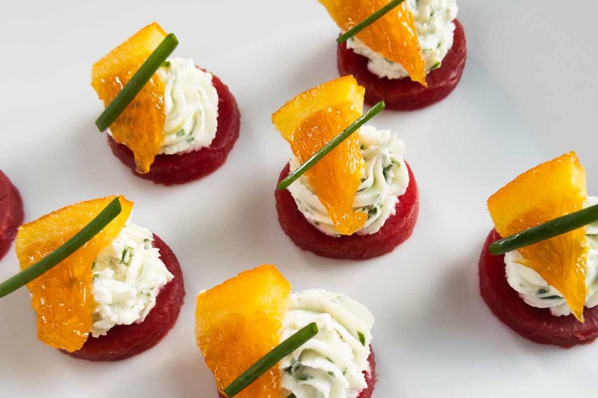 Crowd-pleasing appetizers featuring a red base, piped white cream, an orange slice, and chive garnish—perfect party food arranged on a white surface.