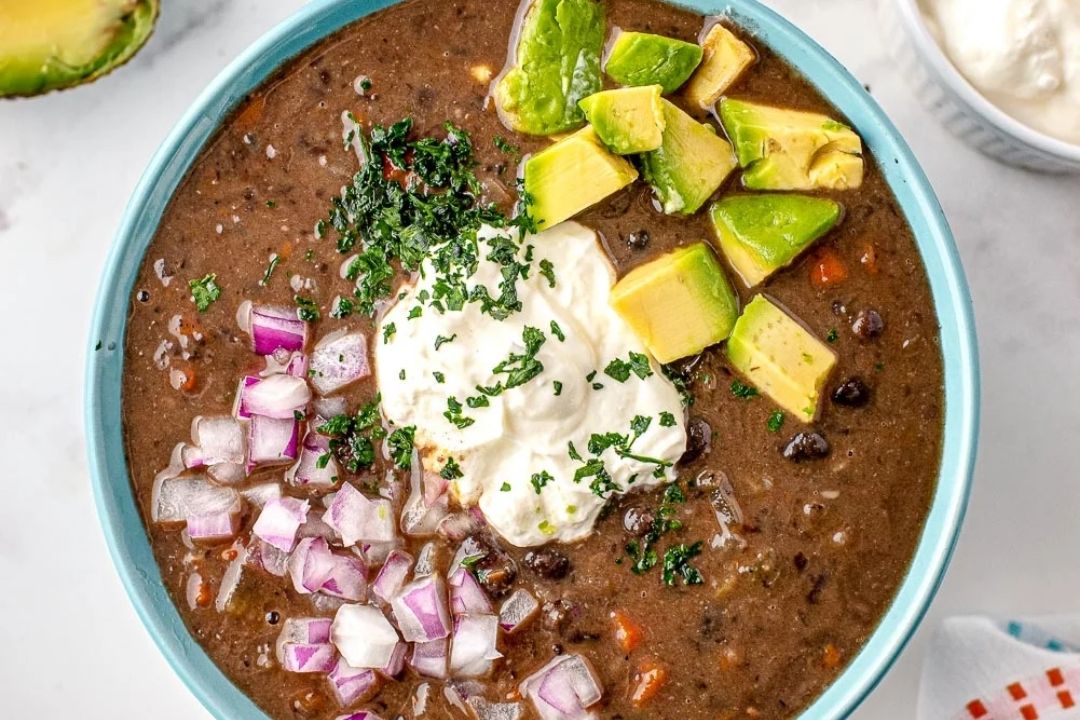 Bowl of black bean soup topped with diced avocado, chopped red onion, sour cream, and herbs, with a small bowl of sour cream in the background—one of the most comforting soups and a delicious addition to your favorite bean soup recipes.