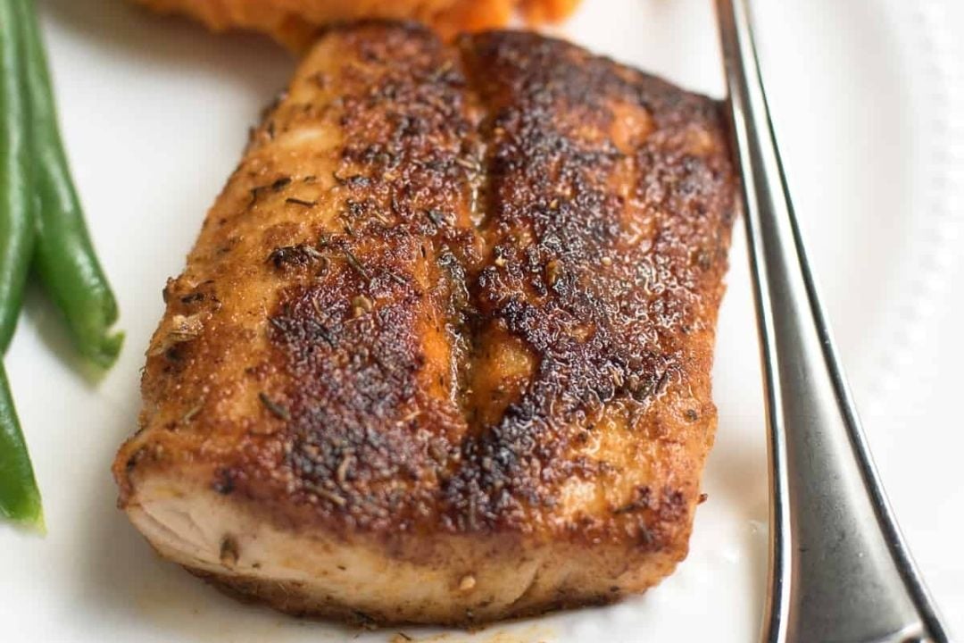 A close-up of a seasoned and seared fillet of fish on a white plate, with green beans and a fork visible beside it—perfect inspiration for those seeking healthy fish recipes.