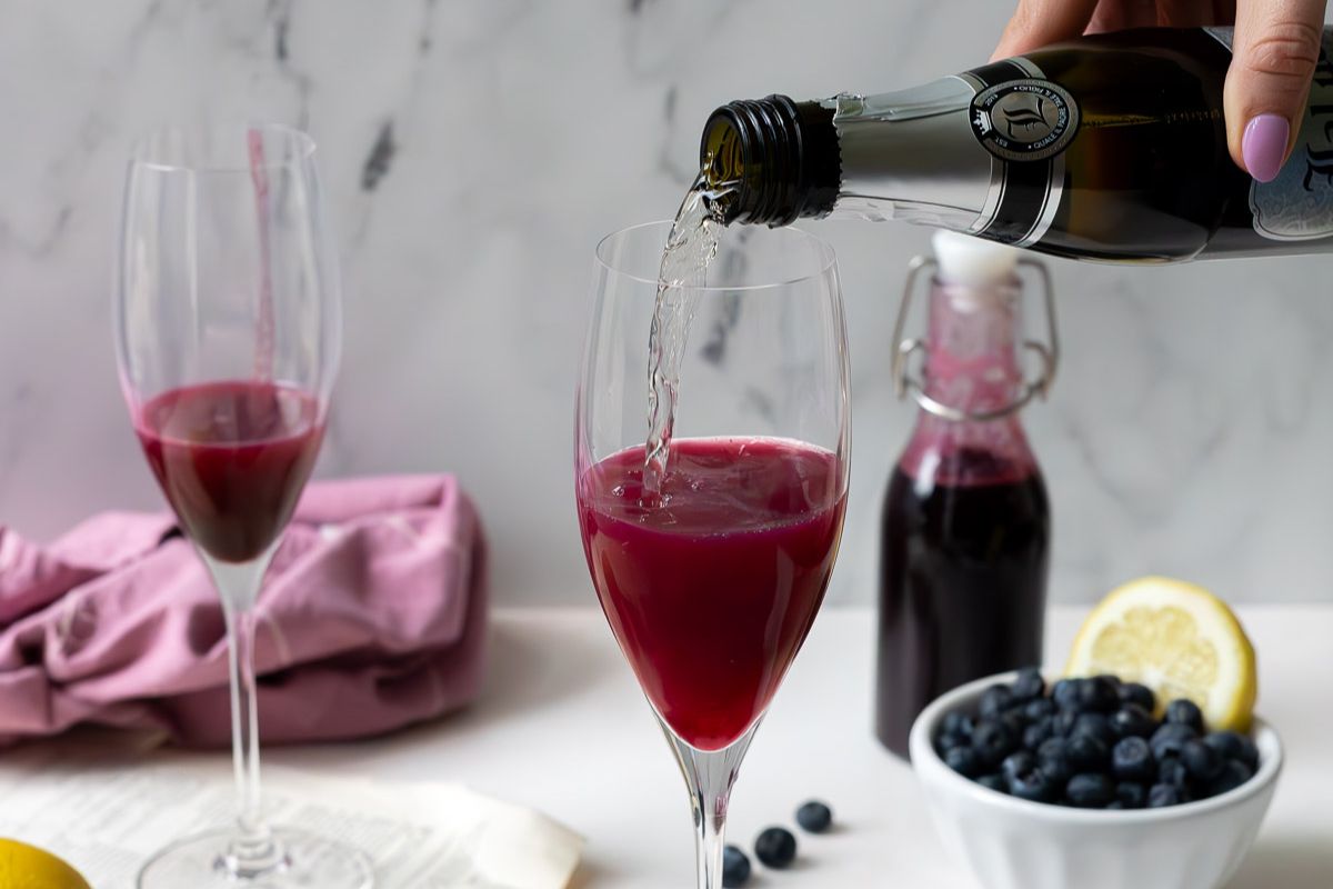 A hand pours clear sparkling wine into a glass of purple juice, creating a refreshing option perfect for brunch cocktails. Nearby on a white surface are another filled glass, blueberries, a lemon slice, and a glass bottle—ideal ingredients for inventive brunch drinks or new cocktail recipes.