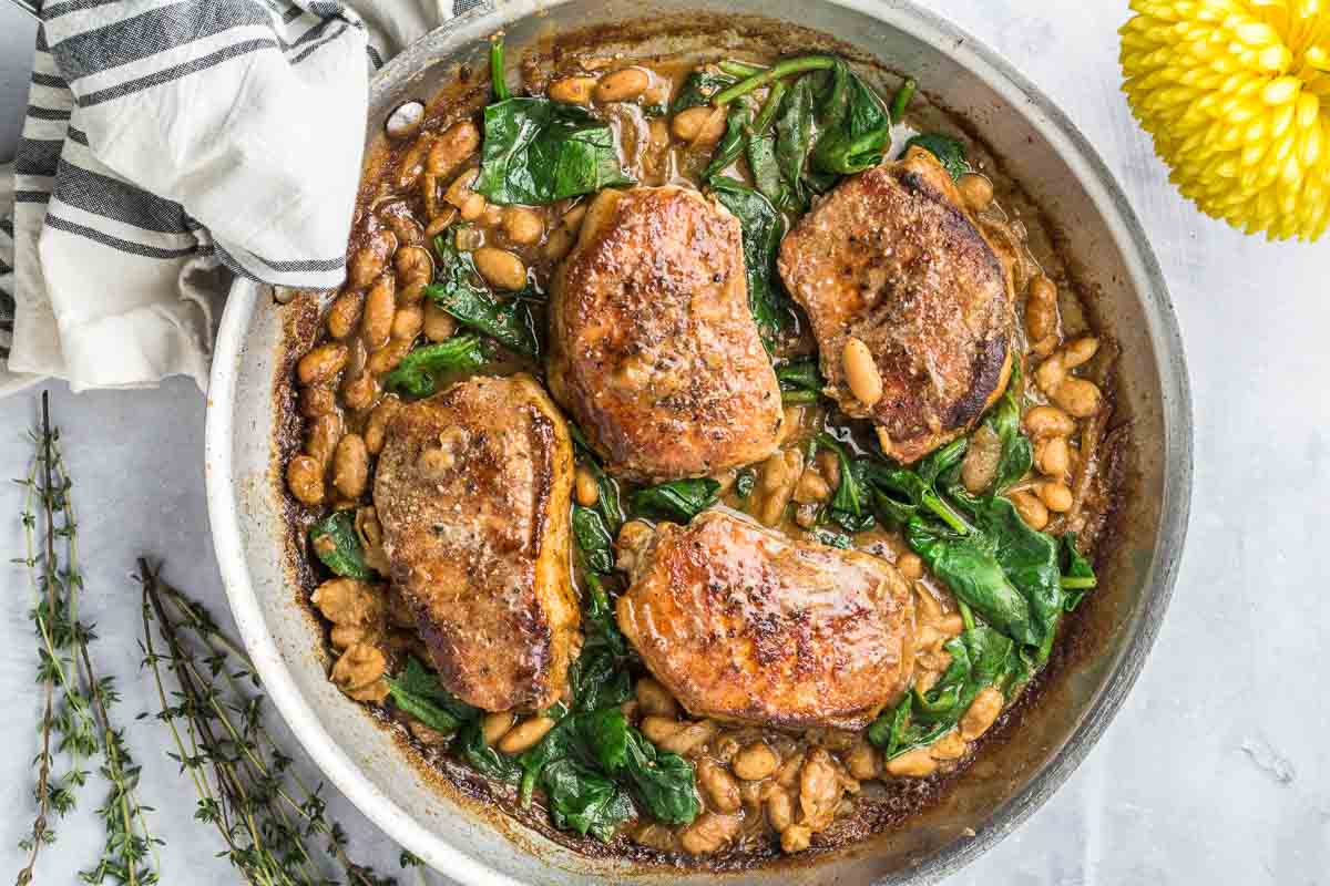 Four cooked pork chops sit in a skillet with white beans and wilted spinach in a flavor-packed brown sauce, making this the perfect dinner recipe. A striped napkin and yellow flower are visible nearby.