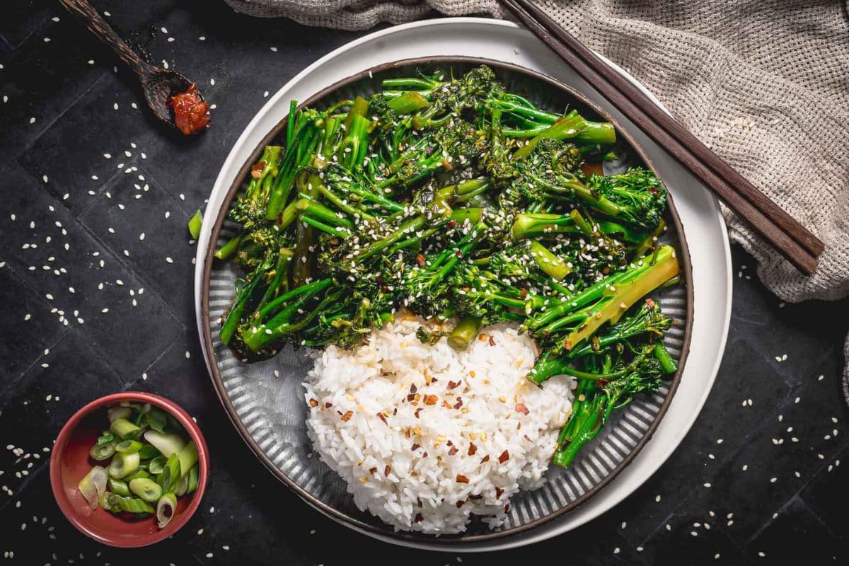 A plate with steamed white rice and sautéed broccolini, sprinkled with sesame seeds—a perfect option for healthy recipes. Chopsticks, a bowl of sliced green onions, and a cloth napkin are nearby, making this ideal for easy recipes or nutritious family meals.