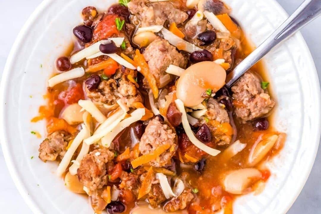 A bowl of sausage and bean stew with tomatoes and shredded cheese sits beside a spoon, making it a perfect addition to your favorite comforting bean soups.