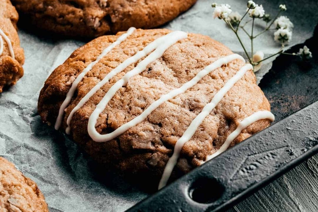 A close-up of a cookie with white icing drizzle on parchment paper, next to a small sprig of white flowers on a baking tray—perfect for easy desserts or as a delightful addition to your favorite autumn desserts.