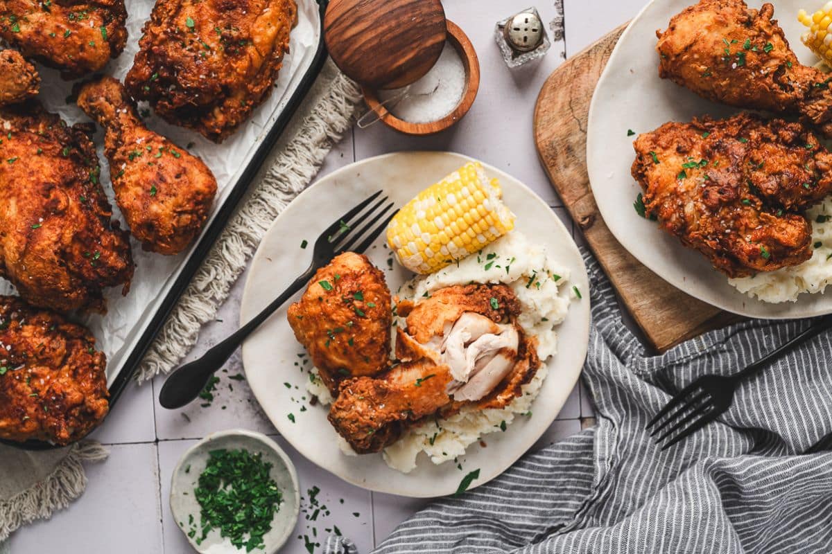 Plates of fried chicken served with mashed potatoes, corn on the cob, and sprinkled herbs, set on a table with utensils and a napkin—classic Southern food that brings the comforting flavors of traditional Southern recipes to your table.