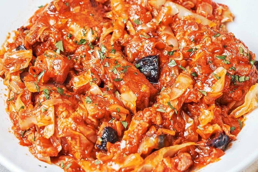 A close-up of a plate of braised cabbage in tomato sauce with herbs and chunks of meat, garnished with parsley—classic comfort food perfect for Sunday dinners.
