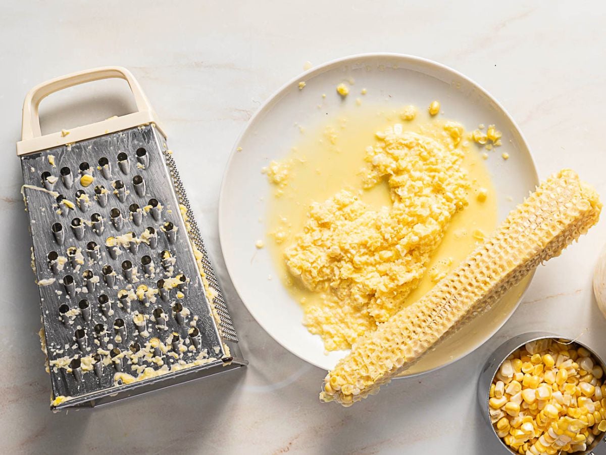 A metal grater, a plate with grated corn and liquid, a stripped corn cob, and a bowl of corn kernels are on a light surface.