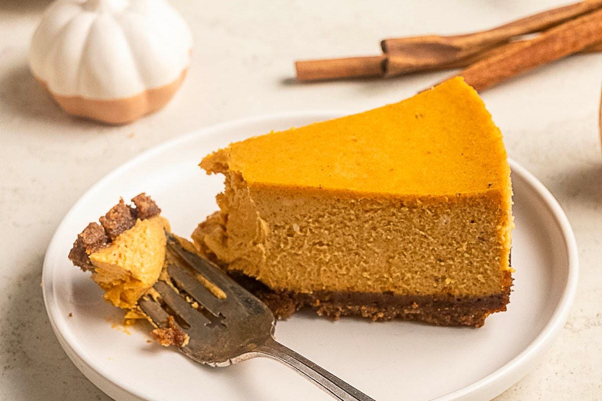 A slice of pumpkin cheesecake with a graham cracker crust is on a white plate, perfect for fans of gluten free desserts. Cinnamon sticks and a ceramic pumpkin are in the background.