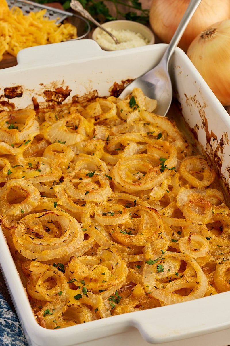 A white baking dish filled with baked cheesy onion rings, garnished with herbs, with a metal serving spoon and shredded cheese and onions visible in the background.