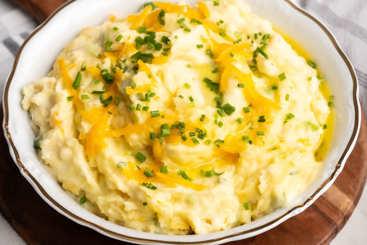 Bowl of mashed potatoes topped with shredded cheddar cheese and chopped chives, placed on a wooden surface—a comforting addition to your collection of German favorites and perfect for serving at any Oktoberfest celebration.