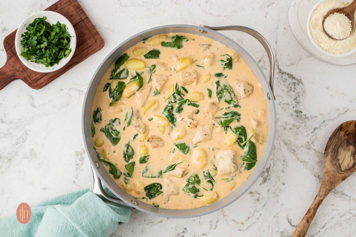 A skillet filled with creamy pasta, spinach, and rotisserie chicken sits on a marble countertop, surrounded by a wooden spoon, chopped herbs, and grated cheese.