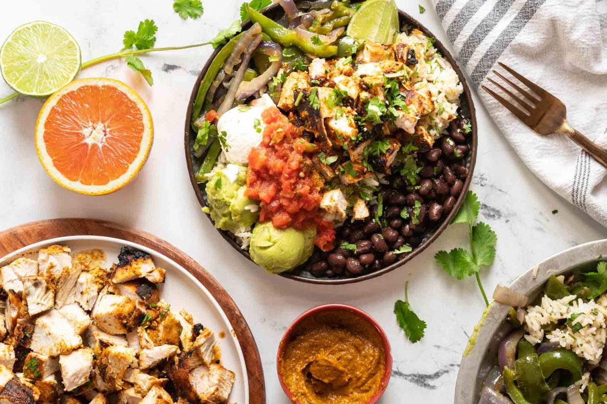 Bowl with grilled chicken, black beans, rice, salsa, guacamole, sour cream, and vegetables on a marble surface—this vibrant dinner is inspired by classic Tex Mex recipes and is surrounded by fresh ingredients and a fork.