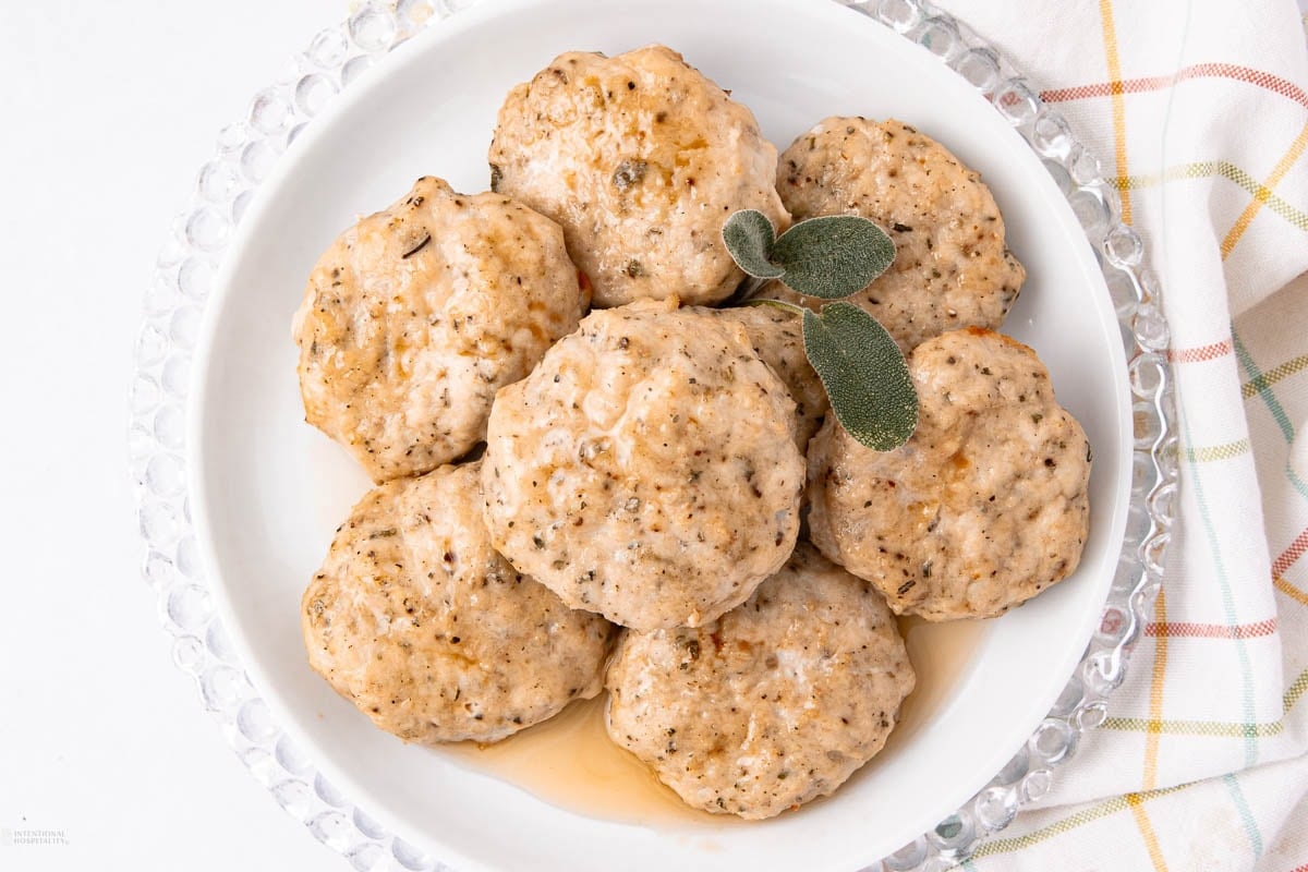 A white plate with seven round, herb-seasoned dumplings topped with a light brown maple syrup sauce and garnished with fresh sage leaves, next to a white and plaid napkin.